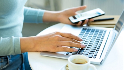 Closeup of a person’s hands – a phone in one hand and the other on a laptop keyboard