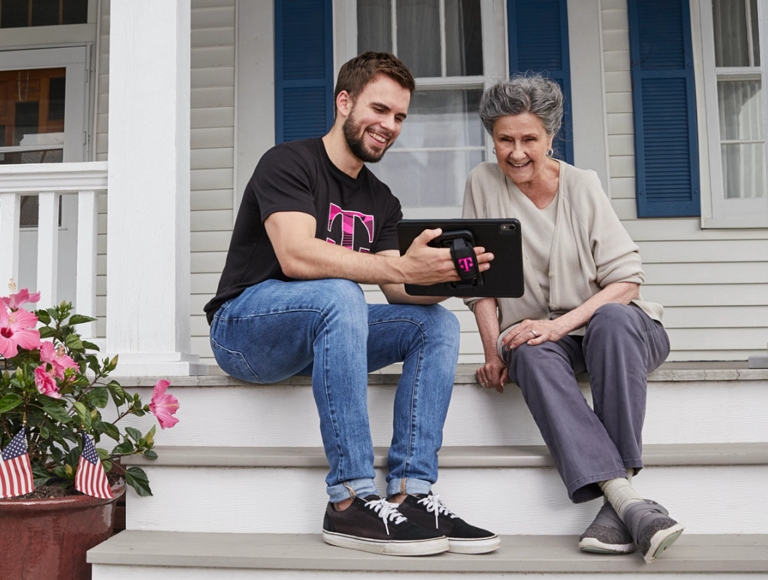 A younger person and an older person sitting on the front steps of a house, sharing a tablet