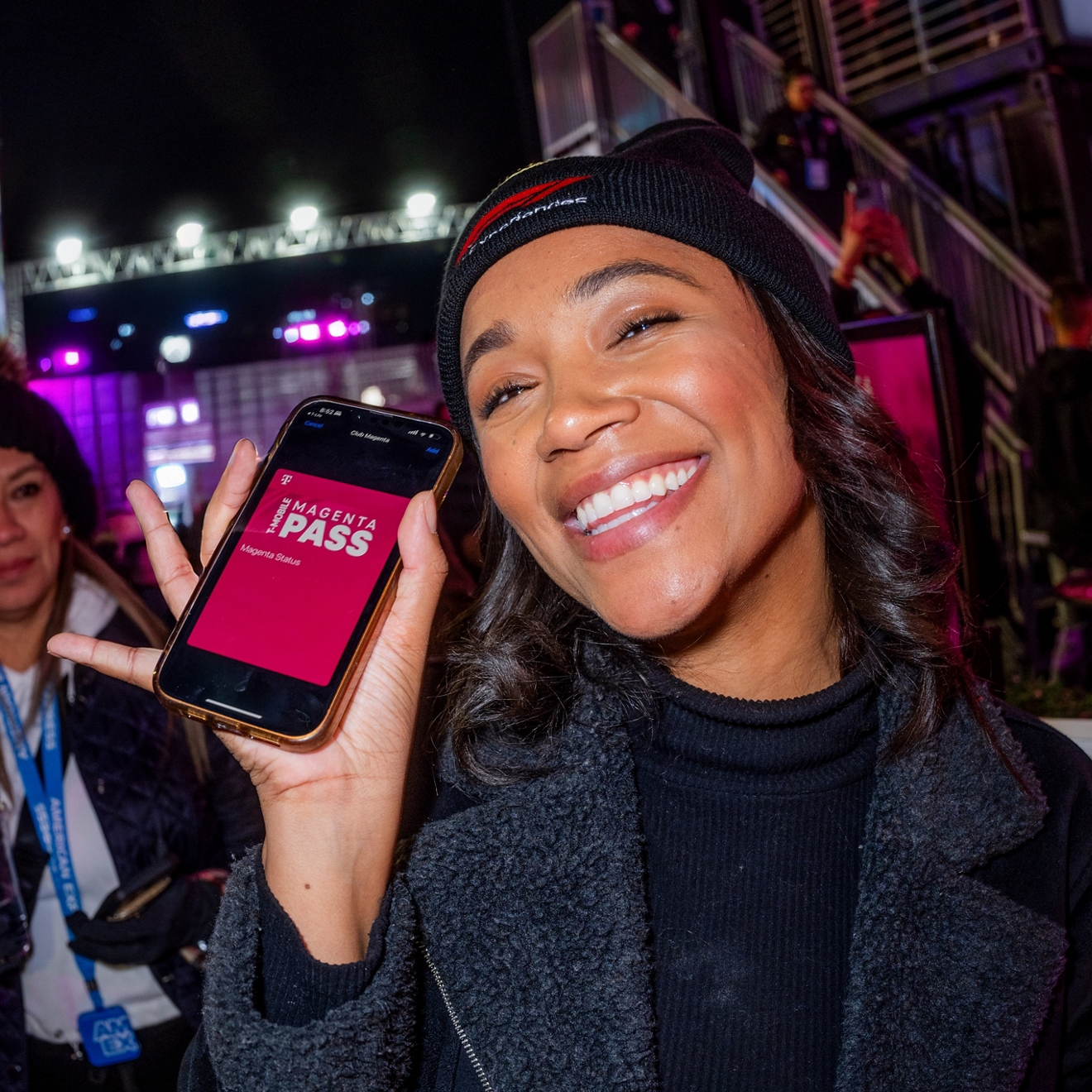 A woman smiling and holding up a phone showing Magenta Pass in T-Life.