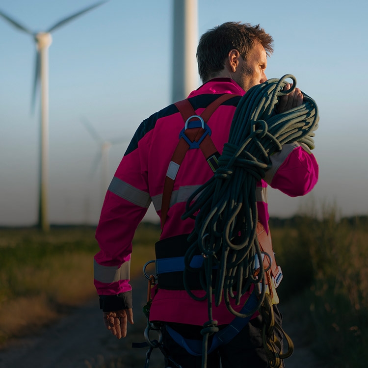A man walks toward a group of windmills. He’s wearing a magenta jumpsuit and carrying rope over his shoulder.