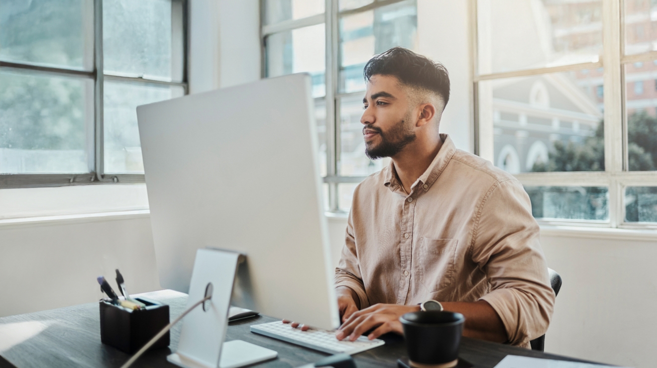 A casually-dressed business professional uses a computer from a sunny, corner office.
