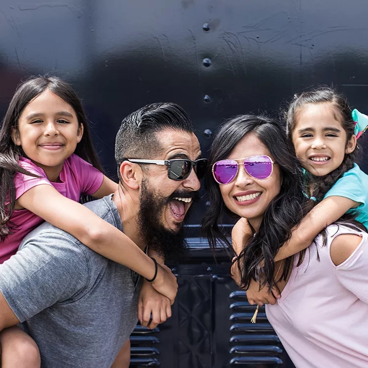 Family of four smiling with one daughter on dad’s back and one daughter on mom’s back.