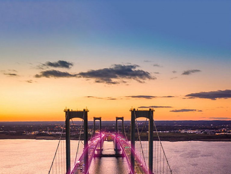 A double suspension bridge spans a body of water, appearing to be lit with magenta beams.