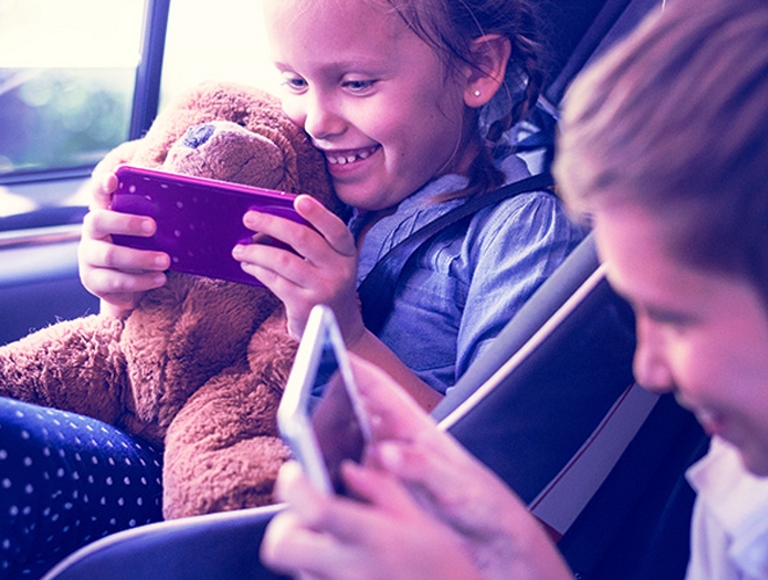 Two children in a car smiling and using smartphones