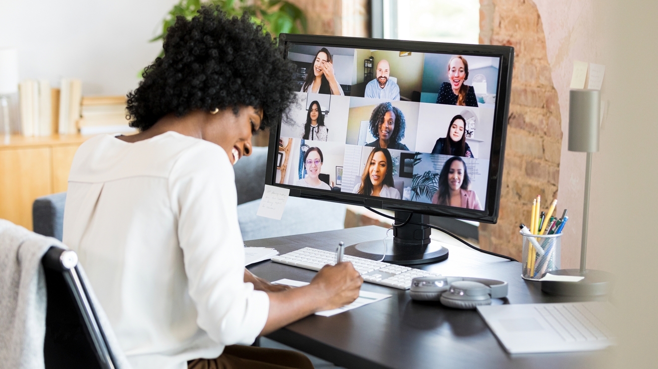 A business professional takes notes at their desk during an online meeting with multiple colleagues.