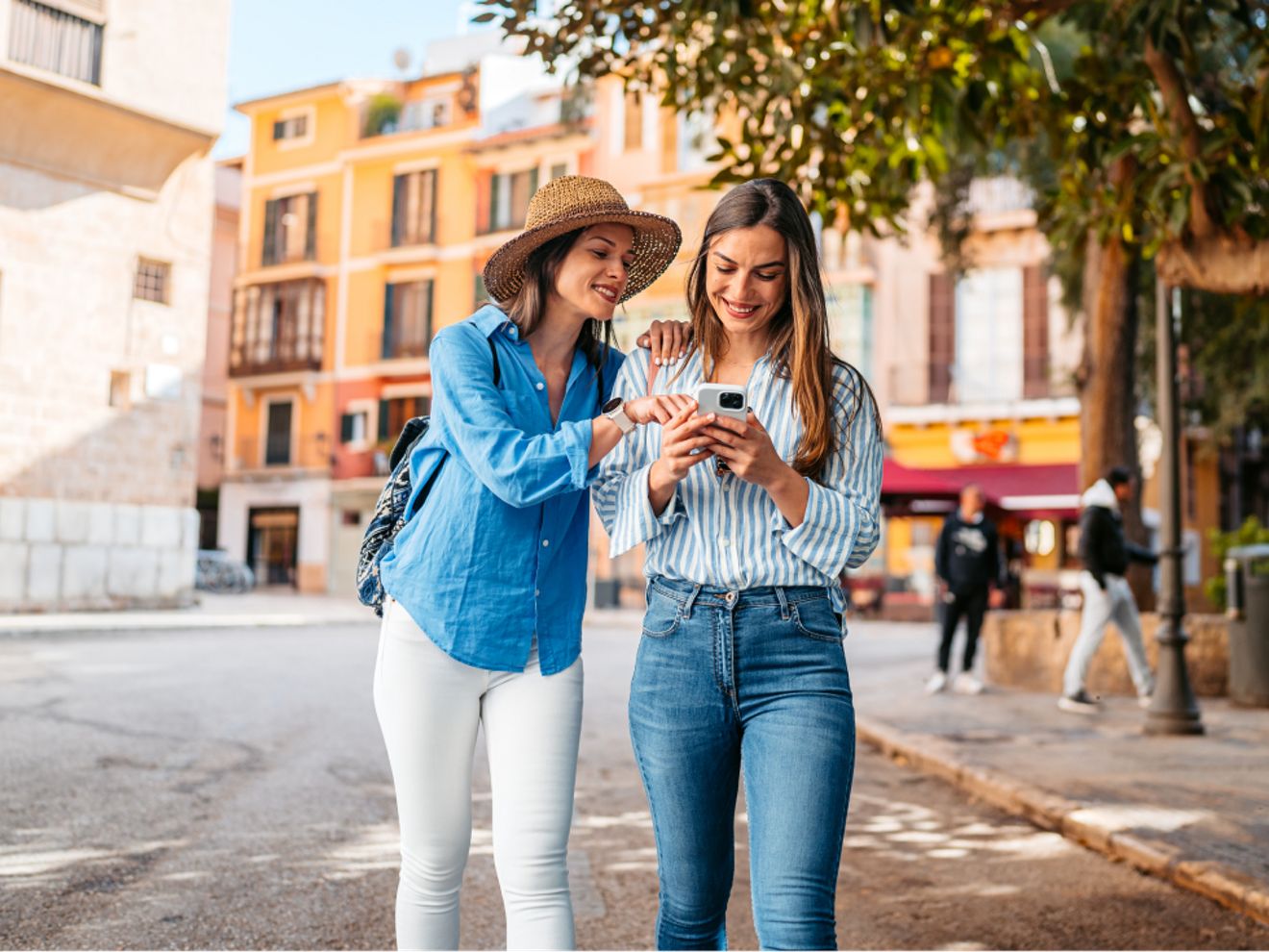 Two women walk together along a sunny city street, smiling as they look at and interact with a smartphone.