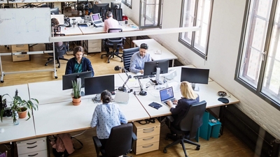 Arial view of a busy, sunlit office filled with employees sitting at desks, working on laptops.