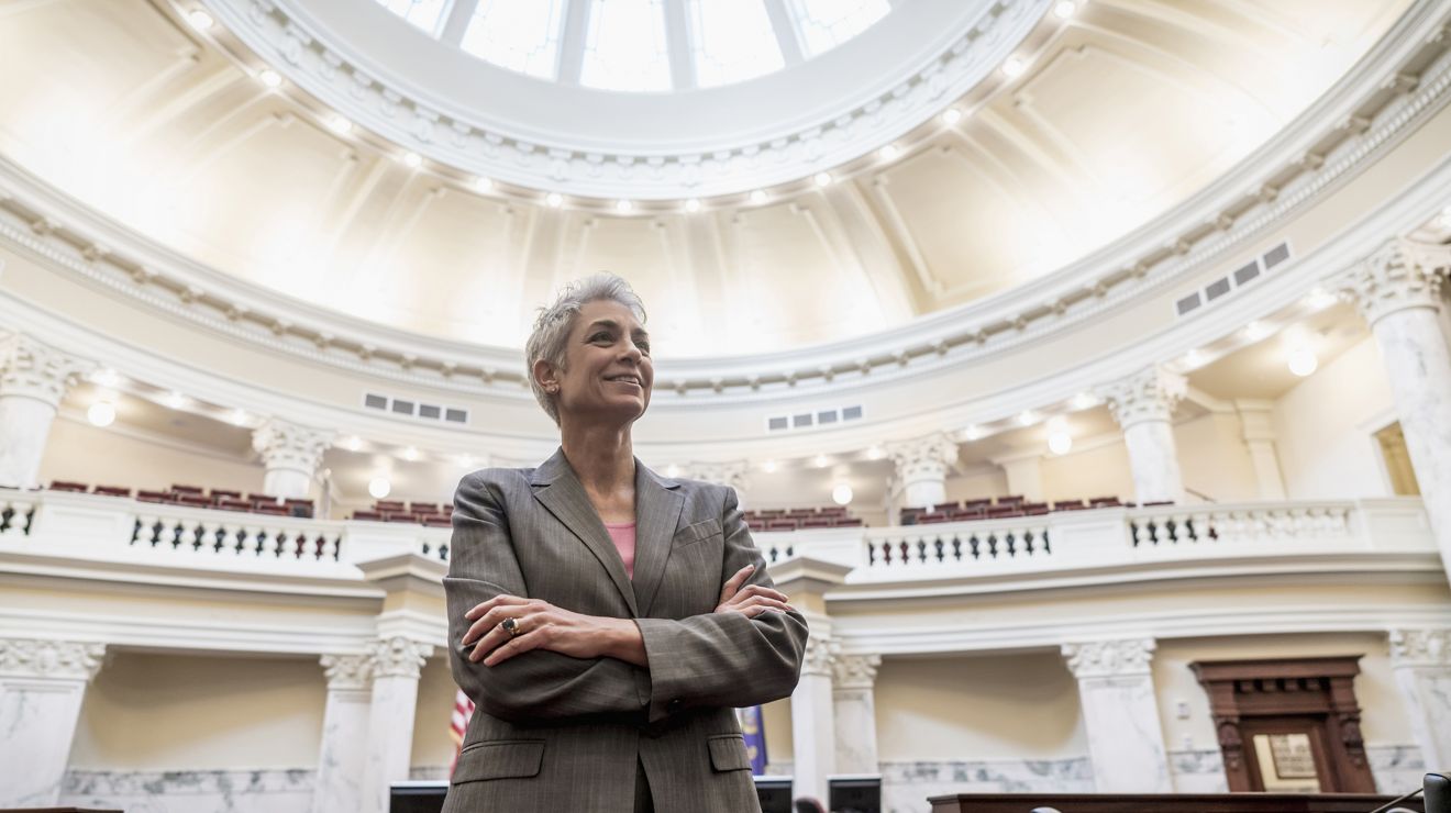 A confident woman in a gray blazer stands with her arms crossed inside a large government  building with a domed ceiling and ornate columns, smiling as she looks slightly upward.