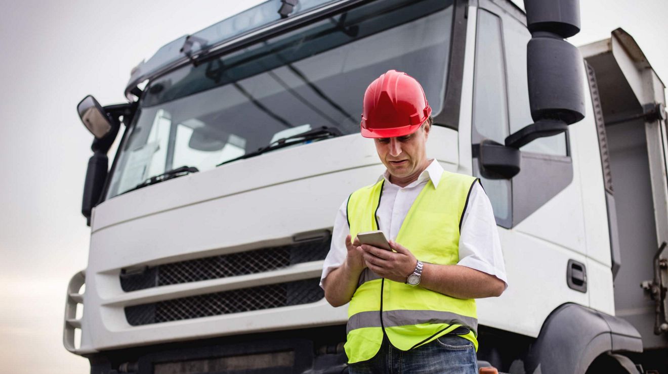 A driver stands beside a semi-truck and uses their smartphone to check in with dispatch.