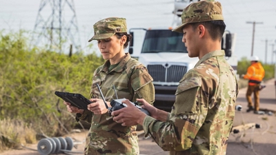 Two uniformed DoD personnel handle a connected tablet and a drone remote control.
