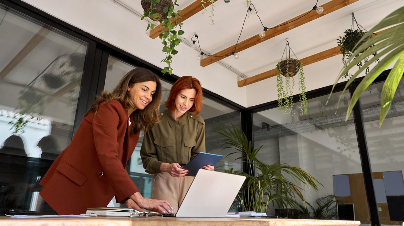 Two women are standing beside a desk in an office, smiling as they look at a laptop. One of the women is holding onto a tablet as she reviews content that’s on the laptop.