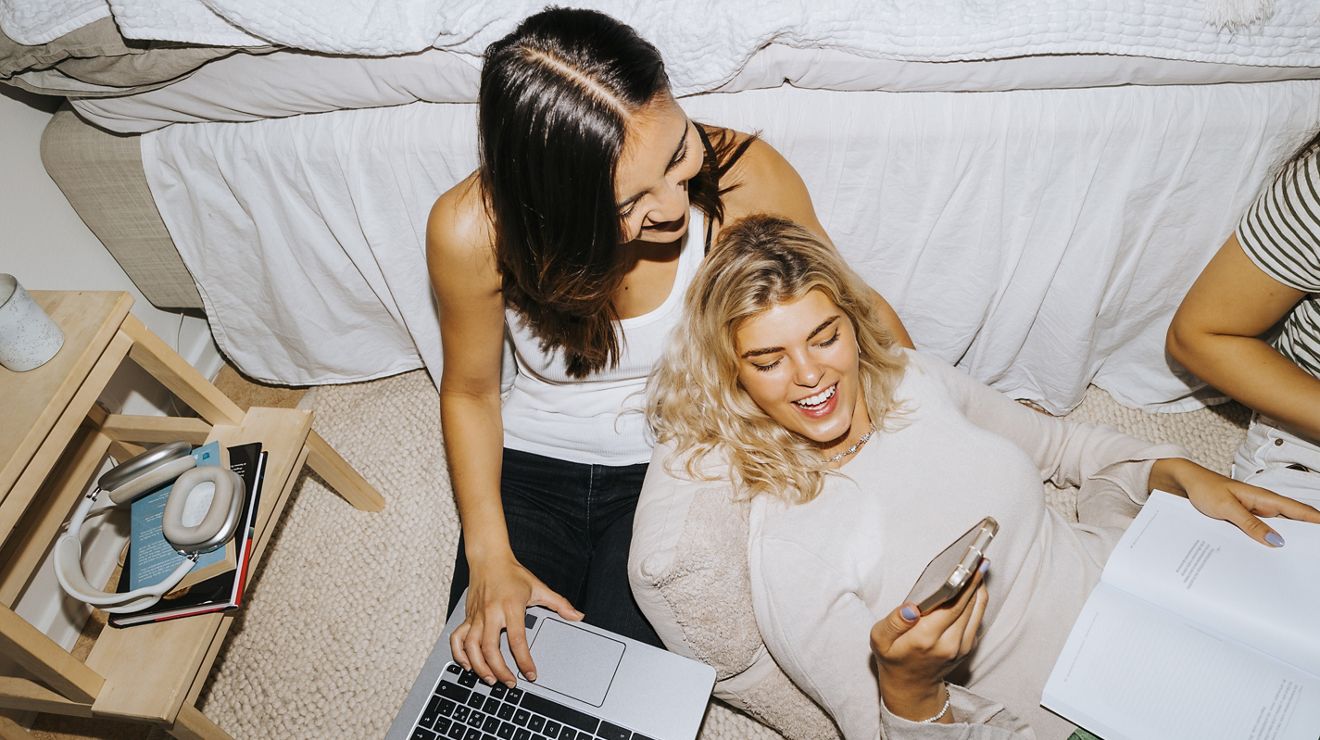 Two women smiling, using a laptop and cellphone, while sitting on a couch