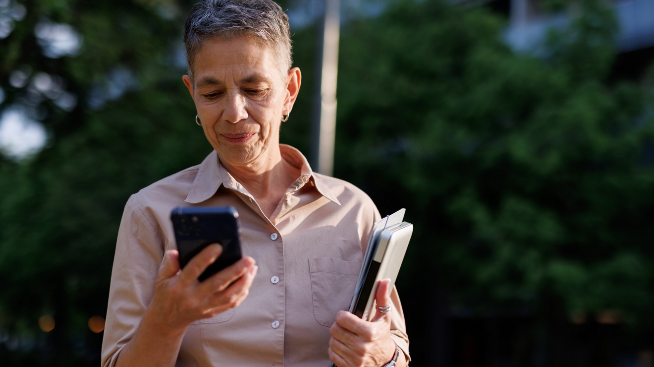 Mature adult professor is smiling while using her phone and holding her laptop