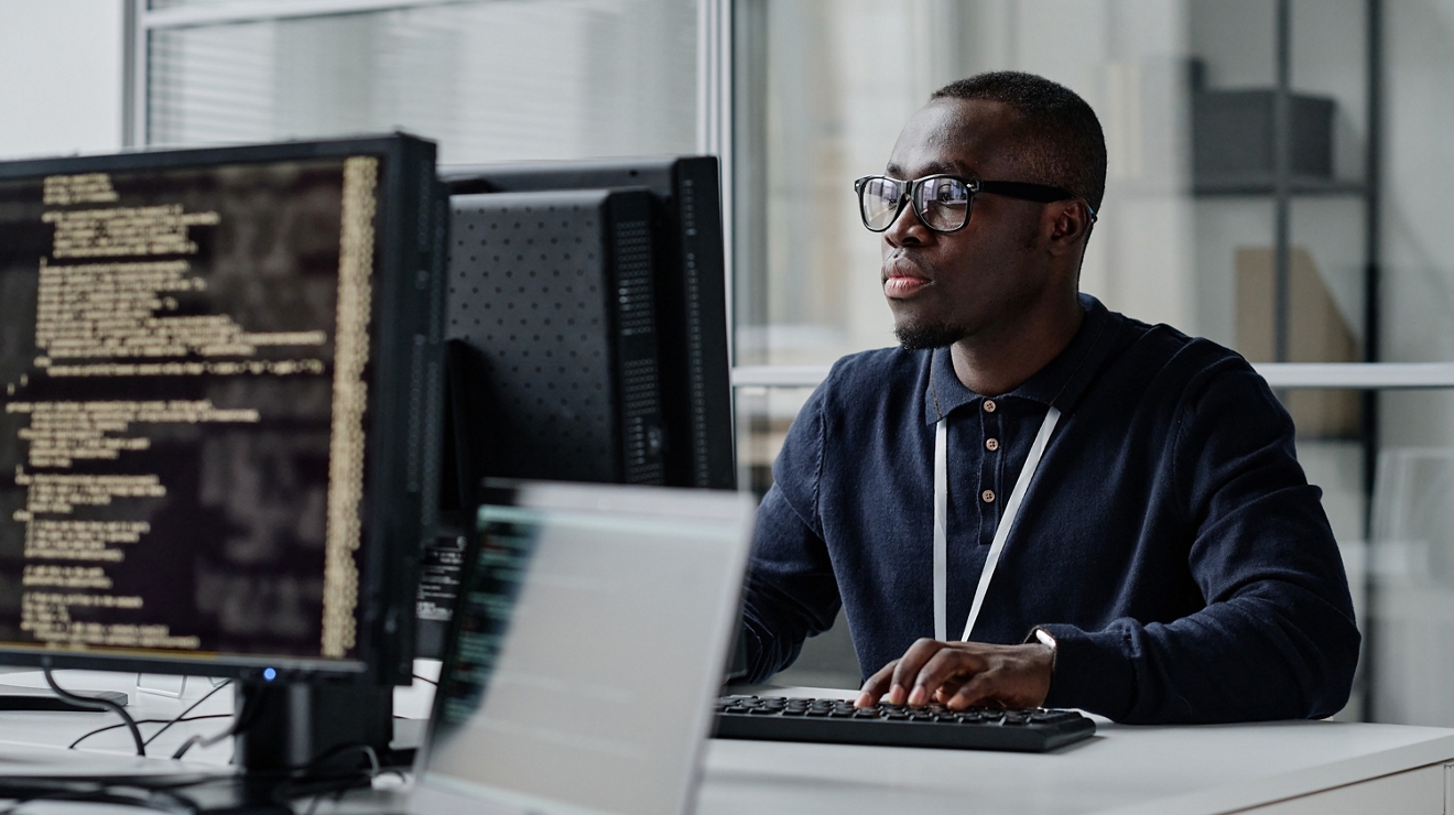 African American young developer in eyeglasses concentrating on his online work on computer sitting at workplace