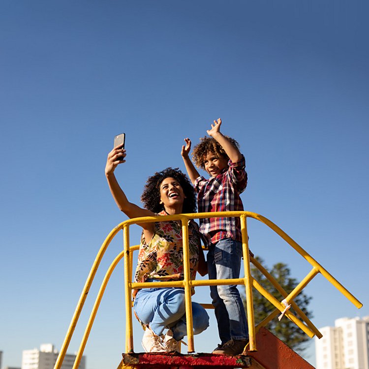 A woman takes a selfie with a child whose arms are up as they stand on top of playground equipment.