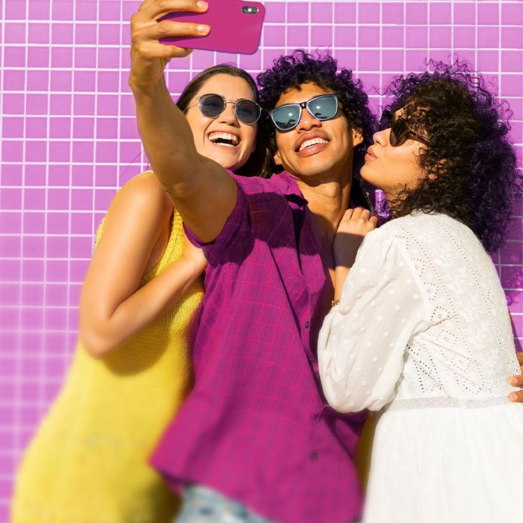 Three friends smile for a selfie while standing in front of a bright magenta background.