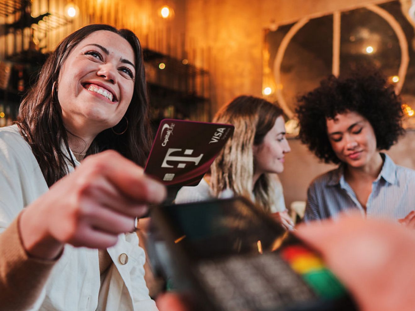 Smiling person taps a credit card to a payment terminal while dining with friends at a restaurant.
