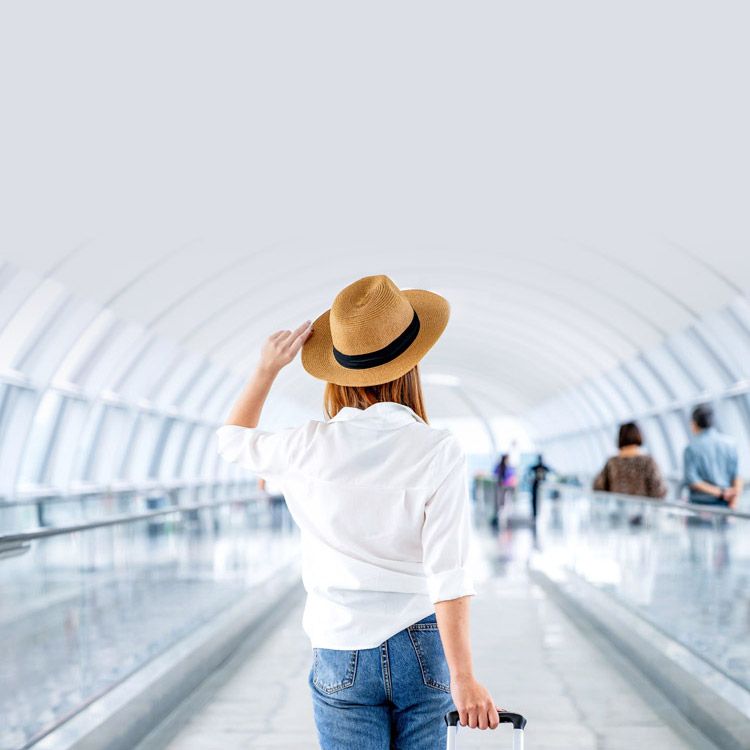 The back of woman in a straw hat rolling her suitcase through an airport walkway, flanked by moving walkways.