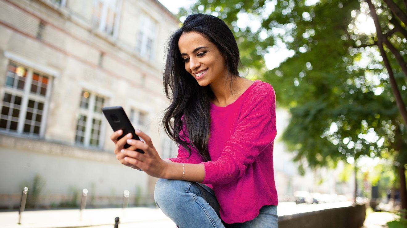 Woman in a pink top smiling while looking at her phone outdoors.