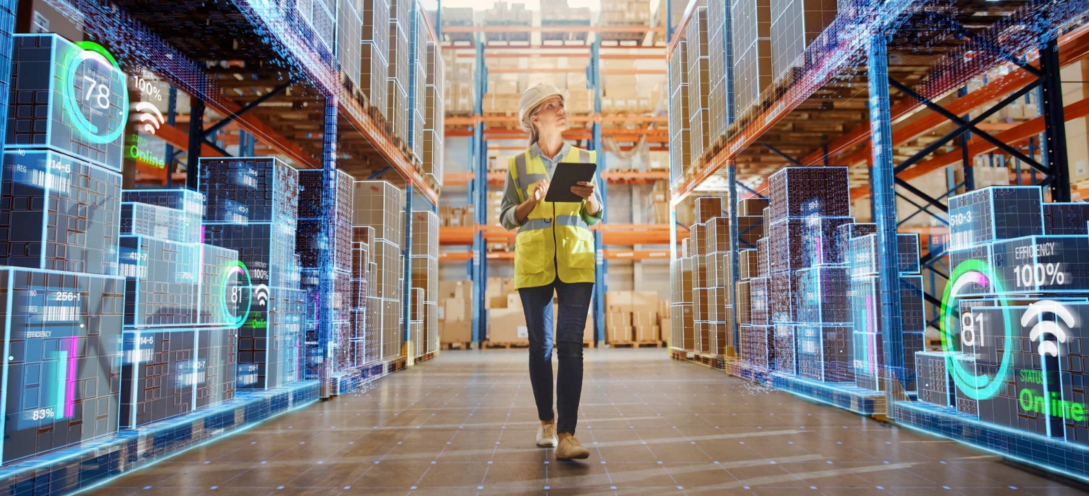 A manager in a hardhat walks a warehouse aisle and checks inventory using a connected tablet.