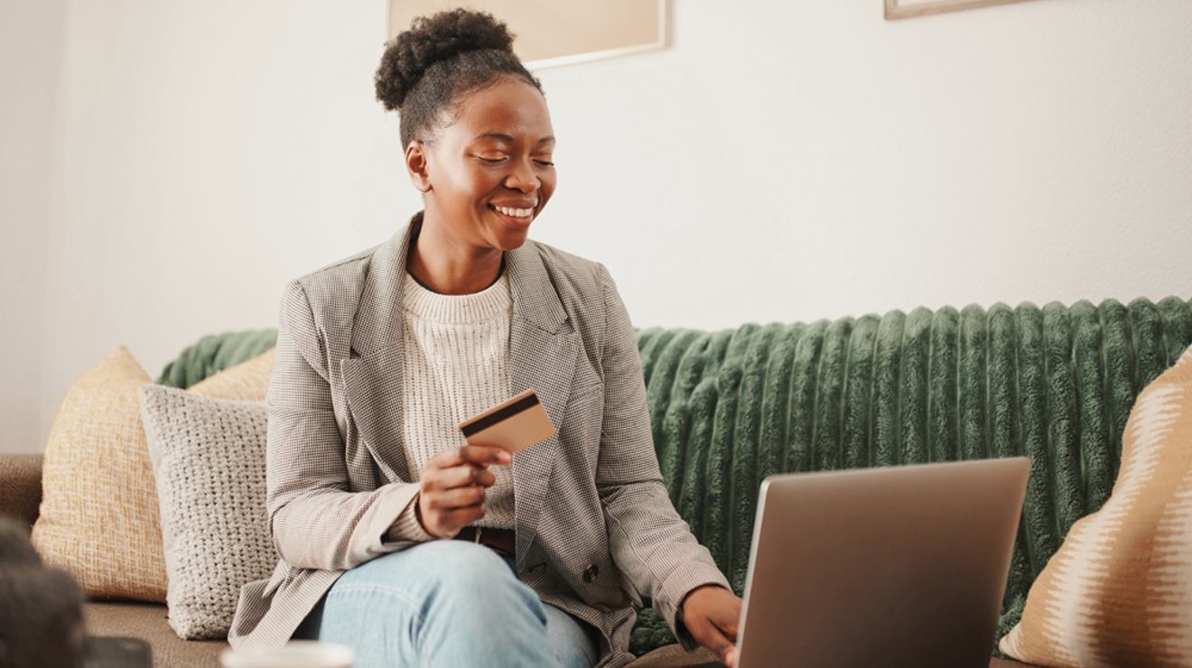 A woman is sitting on a sofa in a living room, smiling as she holds a credit card and types on her laptop.