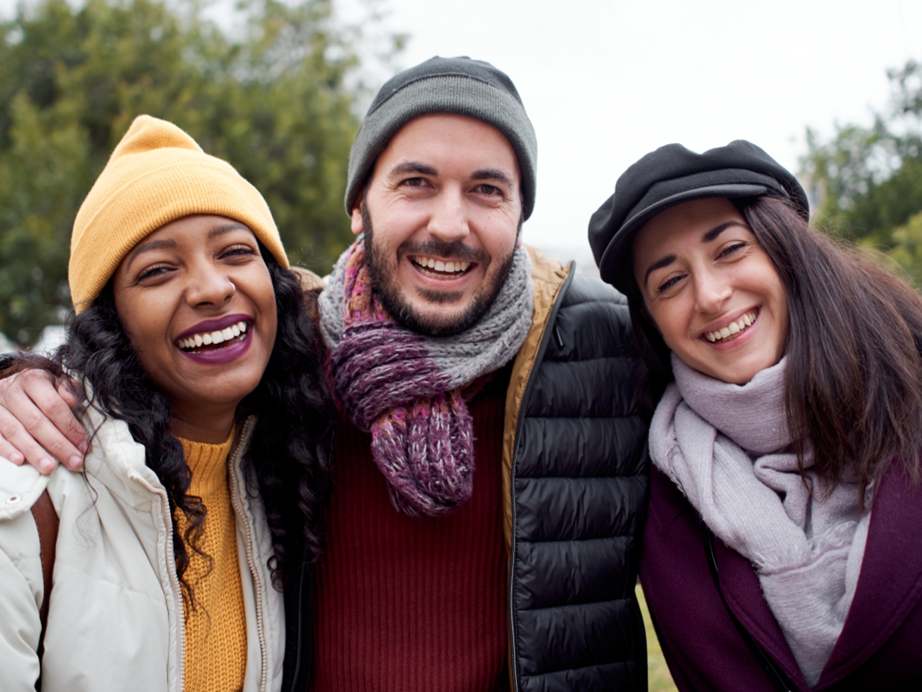 Three friends wearing winter coats and hats smile arm in arm outside.