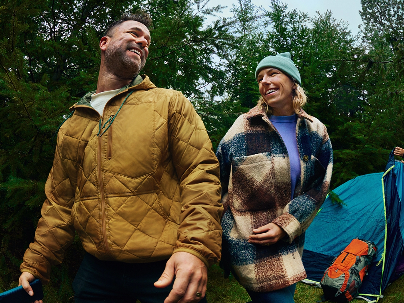 A man holding a cell phone smiles at his friend at a campsite.