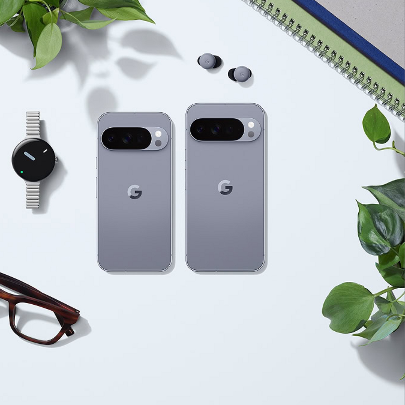 To compare sizes, a Moonstone Google Pixel 10 Pro and Moonstone Google Pixel 10 Pro XL lay side by side screen side down on a flat surface surrounded by plants, glasses, notebooks, earbuds, and a watch. 