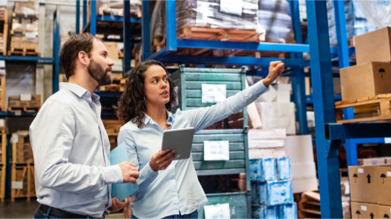 Two warehouse workers gesture to shelves while one holds a tablet.