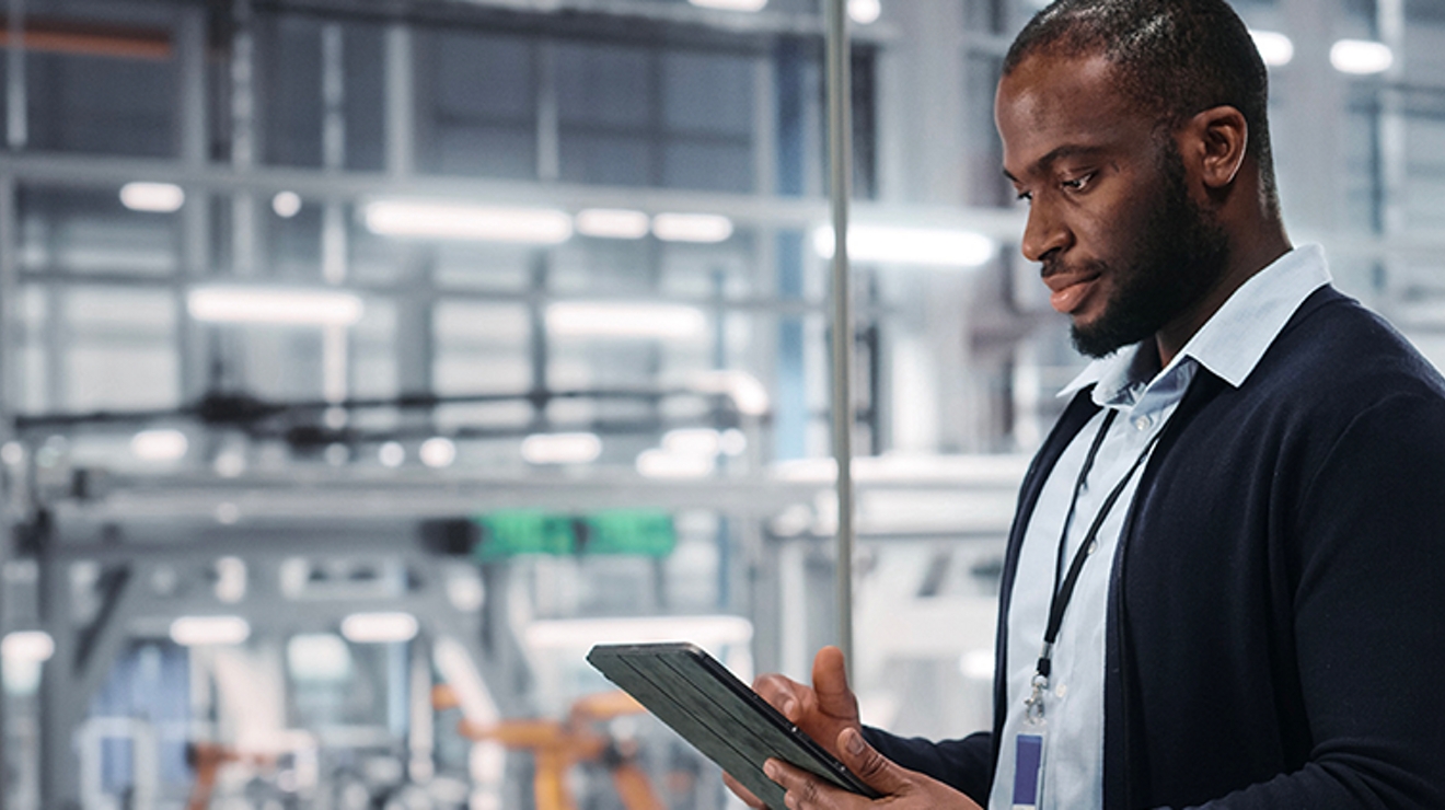 A young professional works on a tablet. A production facility is visible through the window behind him.