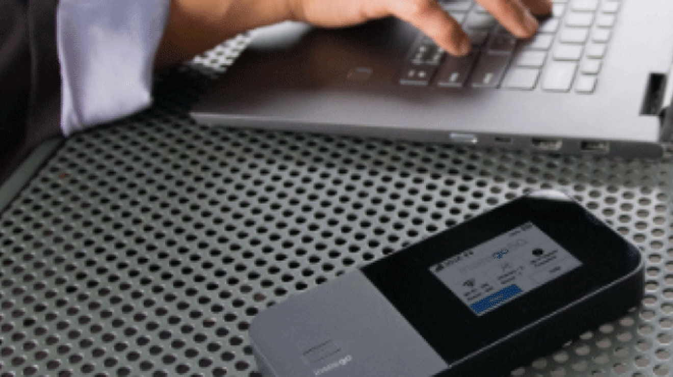 Hands type on a laptop at an outdoor table. A 5G hotspot device resting on the table connects the computer to 5G. 