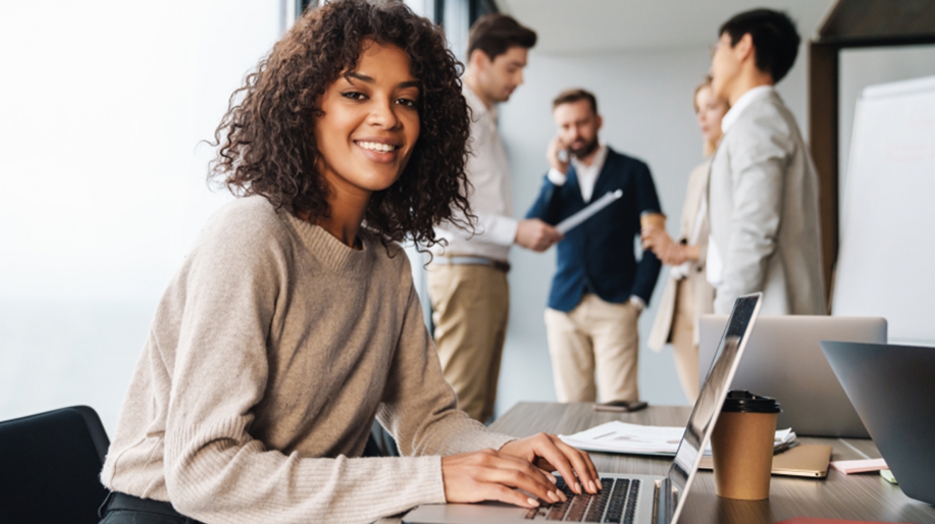 A professional types at her laptop on a conference table while four colleagues collaborate behind her.