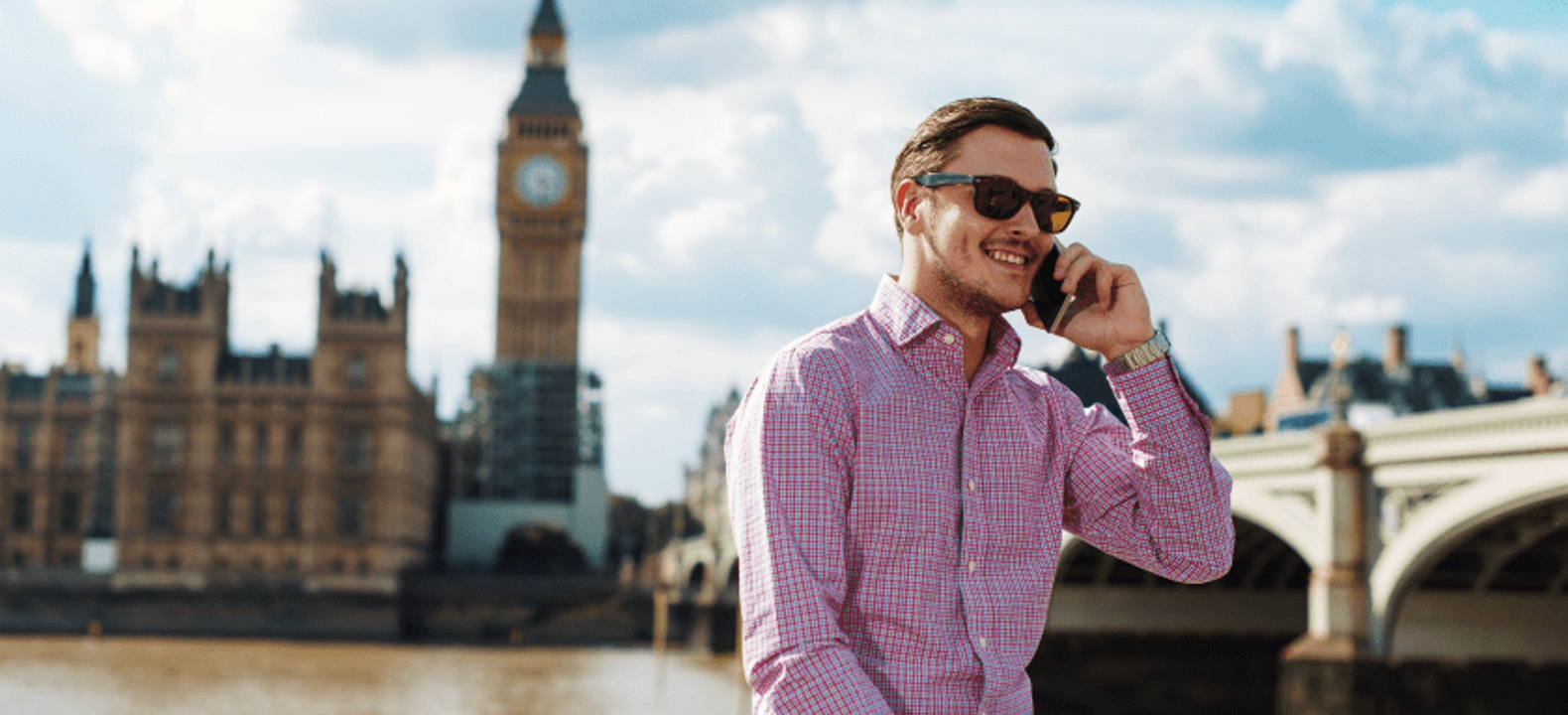 A smiling man takes a business call in London. The Palace of Westminster is visible behind him.