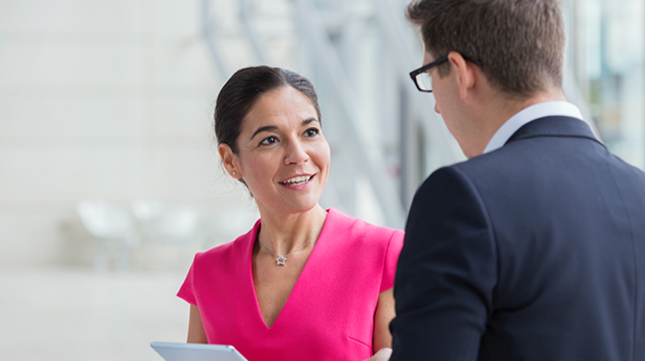 Smiling woman in pink shirt holding tablet while talking to a man in a suit