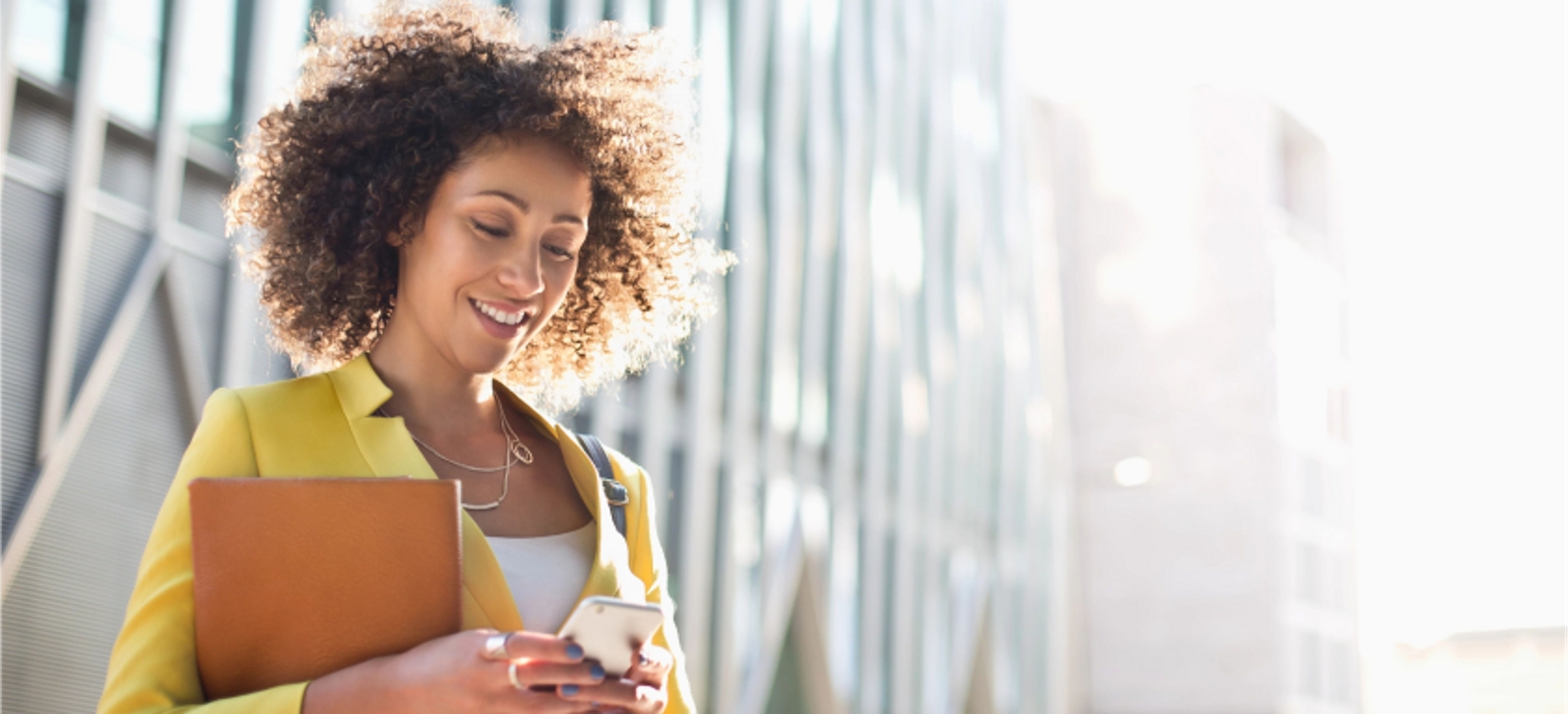 A smiling young professional outside an office tower carries a folder and stays connected on her smartphone.