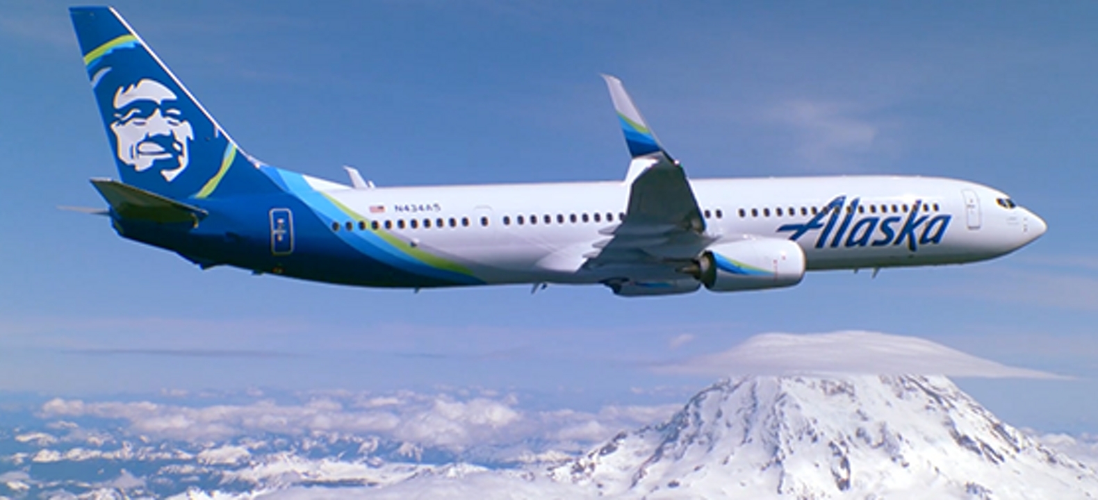 An Alaska Airlines jet flies over the Cascade Mountains in a clear sky.