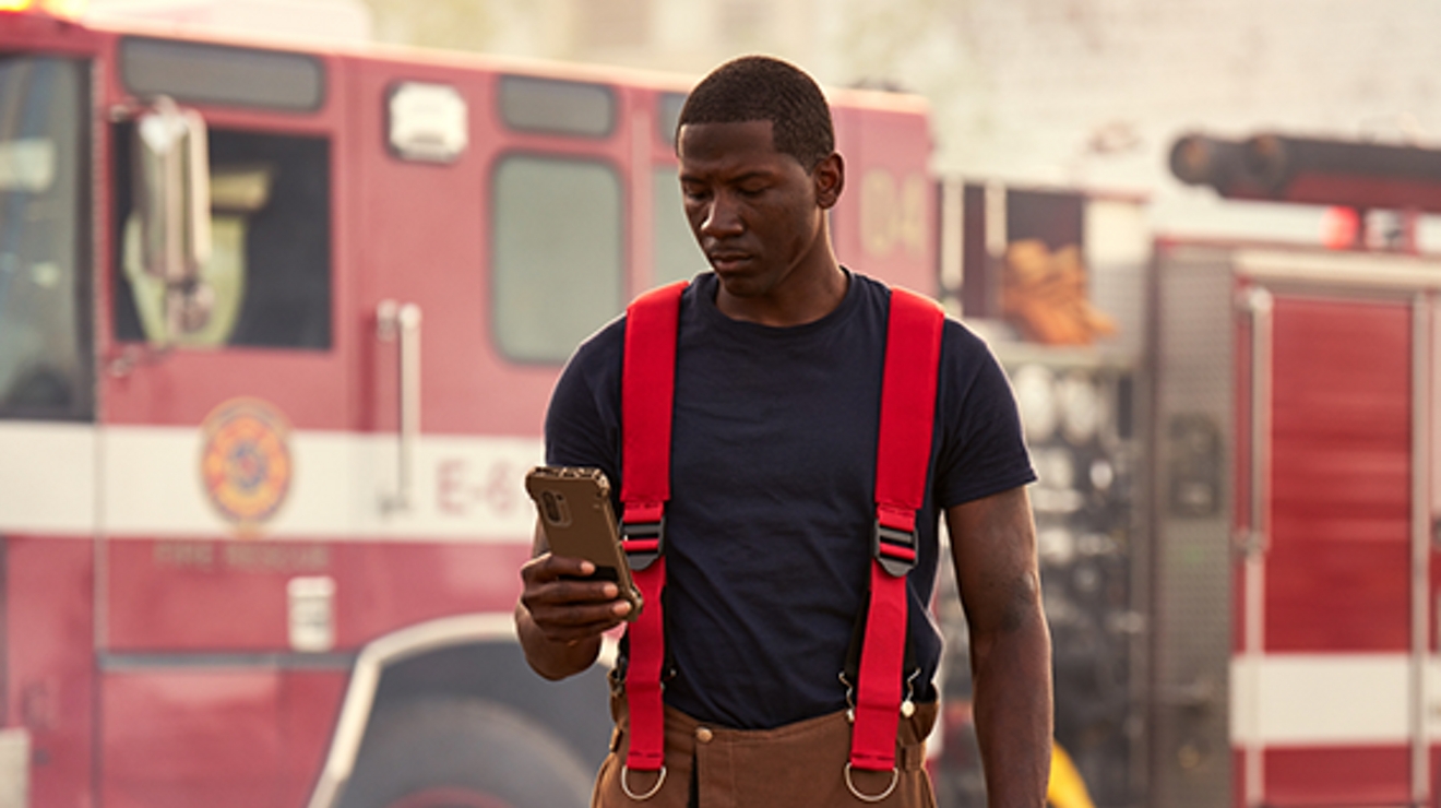 Firefighter in blue shirt with red suspenders stands looking at a smartphone in front of his fire truck