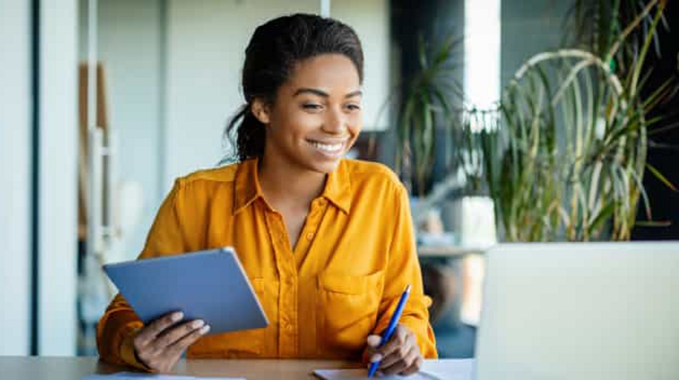 A smiling professional holds a tablet in one hand and takes notes with the other while on a video call on her laptop.