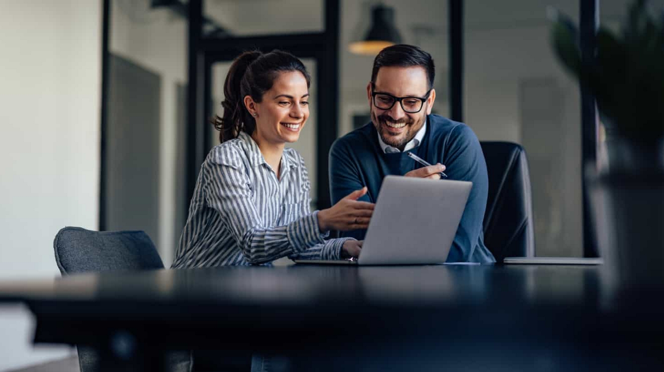 Two colleagues collaborate over a laptop computer while seated in an office.