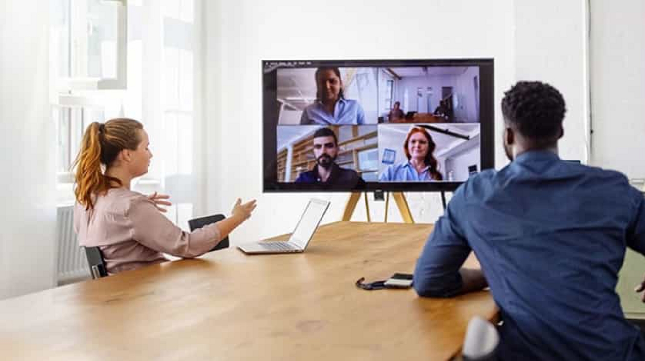 A hybrid meeting with two colleagues in a conference room and three joining on video.