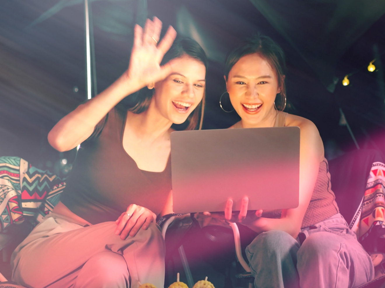 Two female friends sitting outside in the evening, smiling and waving at a laptop during a video call.  