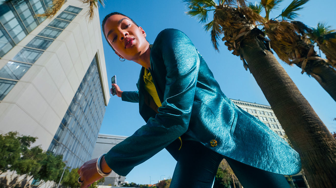 Woman wearing a blue suit looking down as she smirks while buildings and palm trees surround her. 