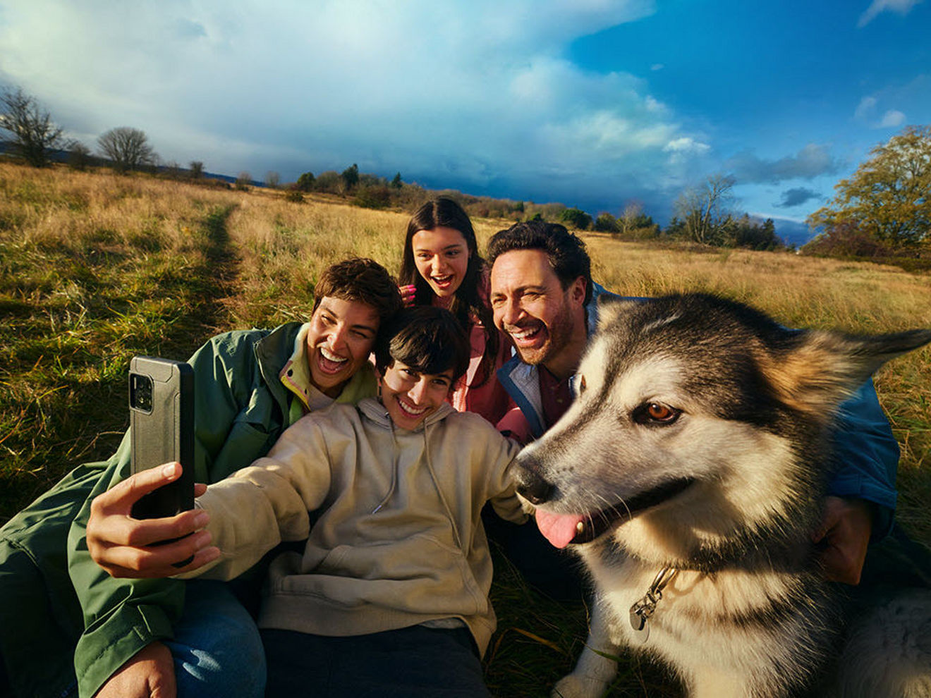 Fall weather outdoors with a man, woman, girl, and boy posing for a selfie with their dog, showcasing friendship and togetherness between adults and children.