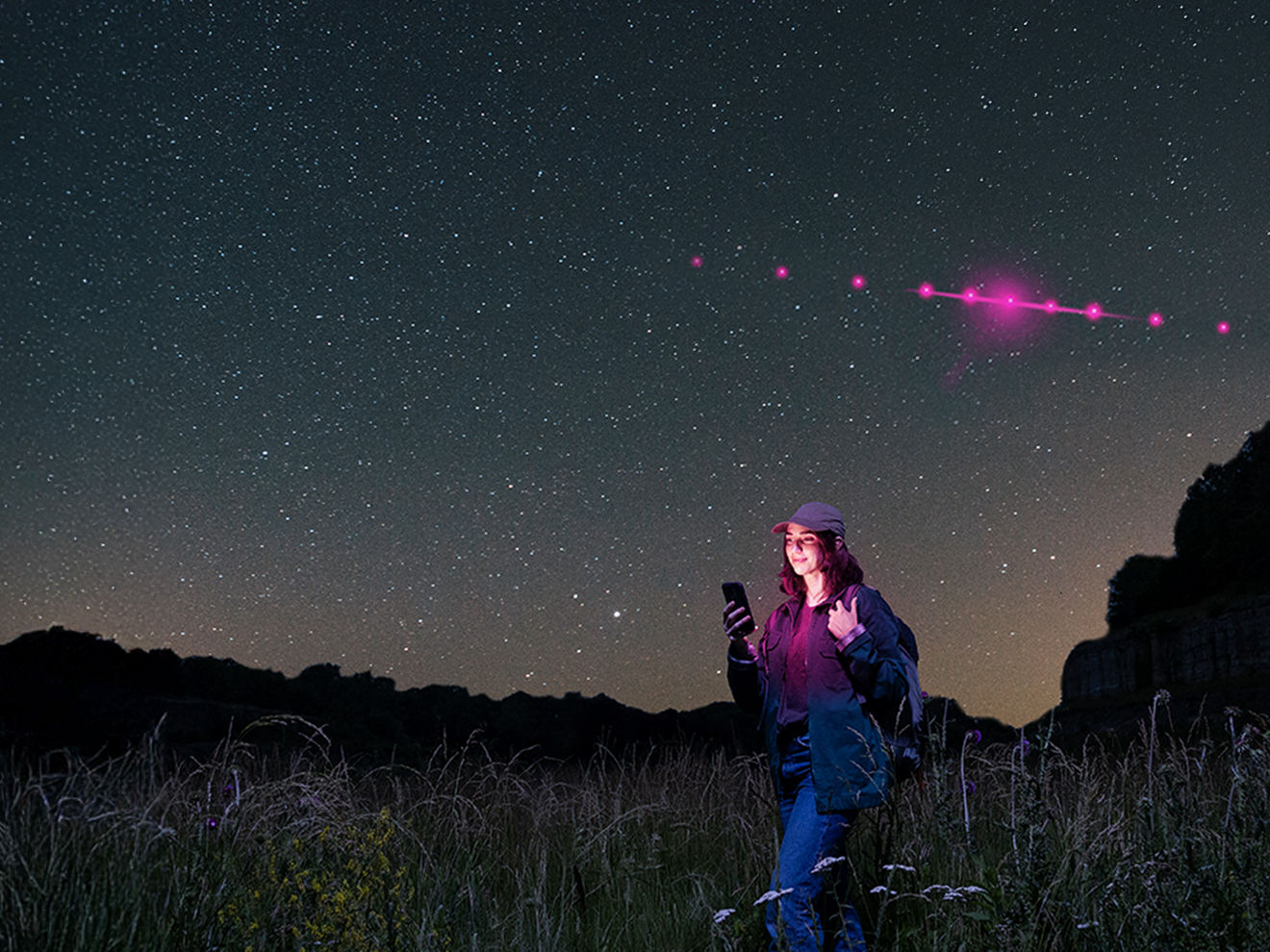 Starry night sky with a woman in a field gazing upward, holding a phone.